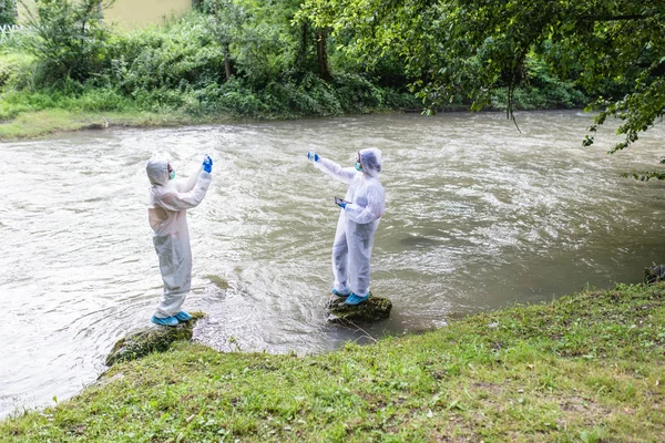 保護スーツを着た2人の科学者が川から水のサンプルを採取 — ストック写真