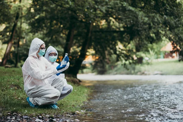 保護スーツを着た2人の科学者が川から水のサンプルを採取 — ストック写真