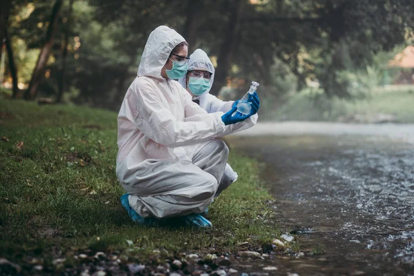 保護スーツを着た2人の科学者が川から水のサンプルを採取 — ストック写真
