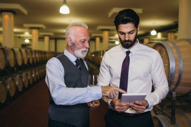 Şarap mahzenindeki şarap üreticileri ellerinde bir kadeh şarap tutup kontrol ediyorlar. Şarap garsonları şaraphanedeki şarapları test ediyor..