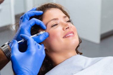 Çekici genç bir kadın canlandırıcı yüz enjeksiyonu yaptırıyor. O, klinikte sakince oturuyor. Uzman güzellik uzmanı hyaluronik asitle kadın kırışıklıklarını dolduruyor..