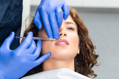 Çekici genç bir kadın canlandırıcı yüz enjeksiyonu yaptırıyor. O, klinikte sakince oturuyor. Uzman güzellik uzmanı hyaluronik asitle kadın kırışıklıklarını dolduruyor..