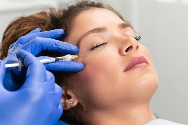 Çekici genç bir kadın canlandırıcı yüz enjeksiyonu yaptırıyor. O, klinikte sakince oturuyor. Uzman güzellik uzmanı hyaluronik asitle kadın kırışıklıklarını dolduruyor..