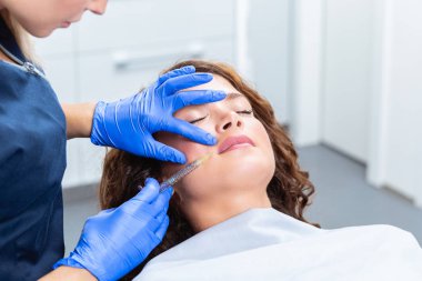 Çekici genç bir kadın canlandırıcı yüz enjeksiyonu yaptırıyor. O, klinikte sakince oturuyor. Uzman güzellik uzmanı hyaluronik asitle kadın kırışıklıklarını dolduruyor..