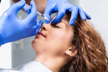 Çekici genç bir kadın canlandırıcı yüz enjeksiyonu yaptırıyor. O, klinikte sakince oturuyor. Uzman güzellik uzmanı hyaluronik asitle kadın kırışıklıklarını dolduruyor..