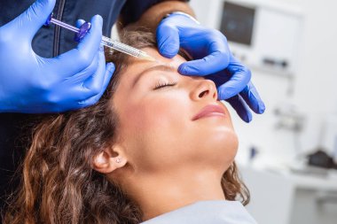 Çekici genç bir kadın canlandırıcı yüz enjeksiyonu yaptırıyor. O, klinikte sakince oturuyor. Uzman güzellik uzmanı hyaluronik asitle kadın kırışıklıklarını dolduruyor..