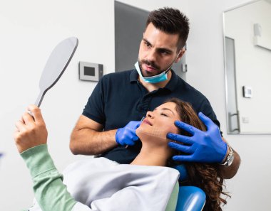 Bir muayene odasında aynaya bakarak yüzünde düzeltme çizgileri ile huzurlu genç hasta.