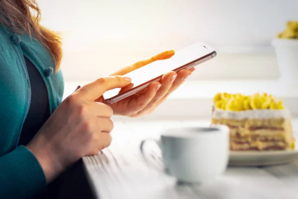 Mobile phone in the hands of a young woman at a table with cake and a cup of coffee