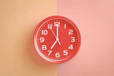 Red clock showing seven o'clock on a beige-pink background clipart
