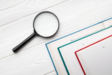 Üst görünüm büyüteç ve beyaz ahşap masa üzerinde farklı renk kapakları açık kitap.