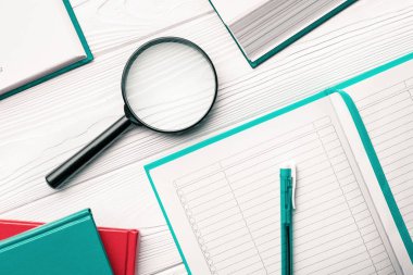 Arama ve araştırma konsepti olarak beyaz ahşap masa üzerinde tükenmez kalem ve kitaplar ile büyüteç ve not defteri