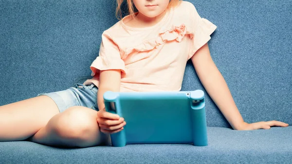 Kanepede uzanmış 5-7 yaşlarında bir kız elinde koruyucu bir çantayla tablet tutuyor..
