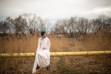 Beyaz uzun bir elbise giymiş bir genç kız, bir boru üzerinde oturan, bir buğday alanı yakın