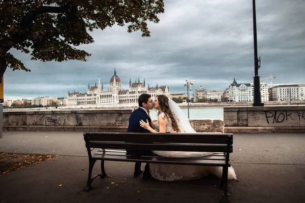 Pengantin wanita dan pengantin pria duduk di bangku dekat sungai di kota tua. Pernikahan di Budapest — Stok Foto