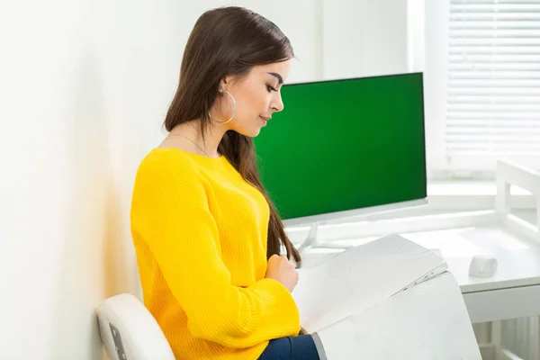 Jonge vrouw die op het werk zit en papier leest. Op de achtergrond is een groen scherm — Stockfoto