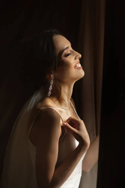 Retrato de novia hermosa en casa. Mujer de pie junto a la ventana en vestido de novia — Foto de Stock