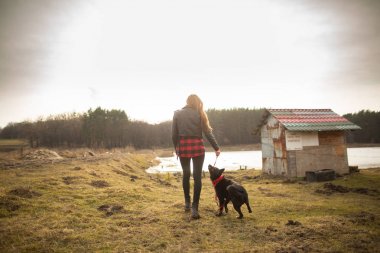 Genç bir kız bir gölün kıyısında köpeğiyle yürüyor. Geri görünüm
