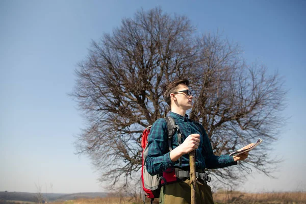 Um viajante com uma mochila, olhando para o mapa e caminhando no campo. Árvore ao fundo — Fotografia de Stock