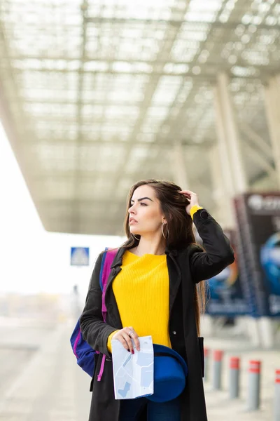 Menina bonita com uma mochila atrás do ombro segurando um mapa, na rua perto do aeroporto — Fotografia de Stock