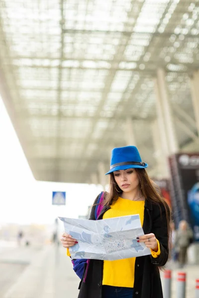 Una joven hermosa con una mochila detrás de sus hombros mira el mapa cerca de un aeropuerto —  Fotos de Stock