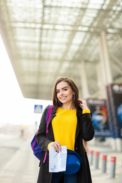 เด็กสาวสวยที่มีกระเป๋าเป้สะพายหลังอยู่หลังไหล่ของเธอถือแผนที่ในถนนใกล้สนามบิน — ภาพถ่ายสต็อก