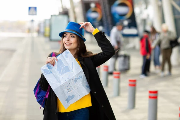 Hermosa joven con una mochila detrás de su hombro sosteniendo un mapa, en la calle cerca del aeropuerto —  Fotos de Stock