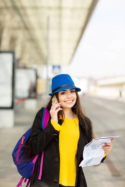 Hermosa joven con una mochila y sombrero azul, hablando en el móvil en la calle cerca del aeropuerto —  Fotos de Stock