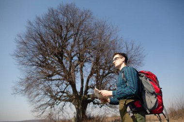 Sırt çantası olan, haritaya bakan ve kırsal kesimde yürüyen bir gezgin. Arka plandaki ağaç