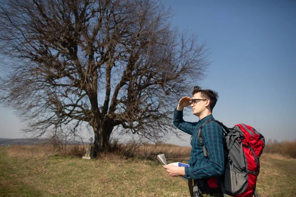 Seorang pelancong dengan ransel, melihat peta dan berjalan di pedesaan. Pohon di latar belakang — Stok Foto