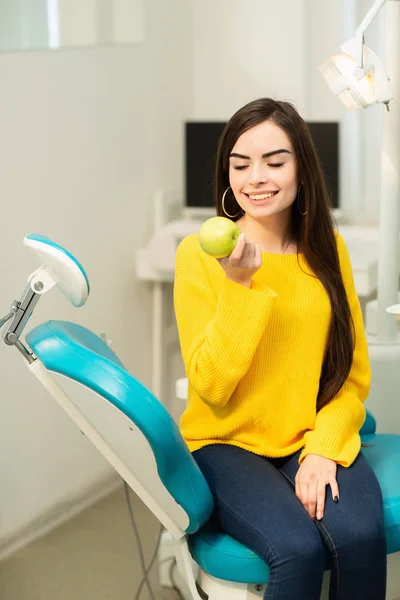 Happy girl sitting in dental chair and showing fresh apples after successful dental treatment — Stock Photo, Image