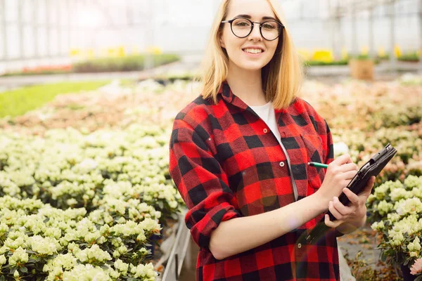 Mujer joven trabajando en invernadero. Muchacha atractiva comprobar y contar las flores, utilizando el ordenador tableta —  Fotos de Stock