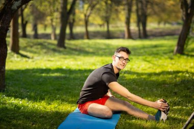Gözlüklü genç adam dışarıda yoga eğitimi alıyor. Sportif adam, parkta, mavi yoga minderinin üzerinde rahatlatıcı egzersiz yapıyor. Boşluğu kopyala