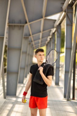 Adam açık havada tenis raketve bir top ile poz veriyor. Omzunda bir havlu asılı. Spor konsepti