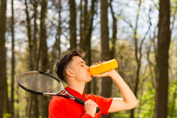 The man in a red t-shirt pose with a tennis racket and drinking water from a thermocouple. Sport concept — Stock Photo, Image