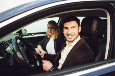 Genç bir adam araba kiralıyor. Satış merkezinin çalışanı arabada belgeler gösteriyor.