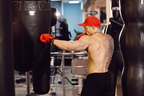 Petinju berotot bertelanjang dada dengan karung tinju di gym. Seorang pria dengan tato di sarung tinju merah . — Stok Foto