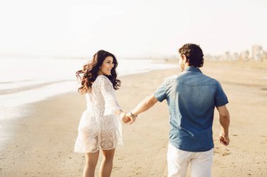 Mutlu çift deniz boyunca yürüyor, el ele tutuşuyor. Beyaz elbiseli kız, kot pantolon giyen adam. Aşk hikayesi. Geri görünüm