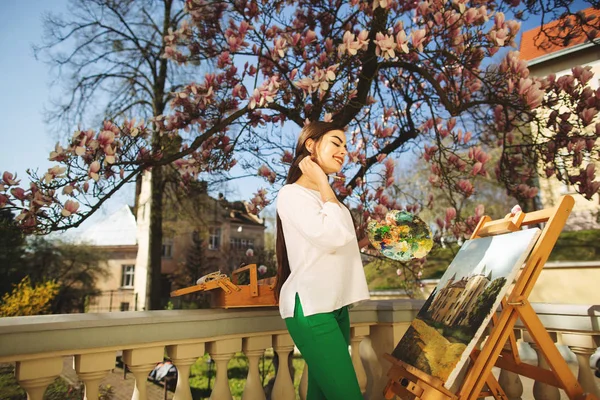 Giovane artista bruna sorridente dipinge un quadro per strada, vicino a un bellissimo albero di magnolia — Foto Stock