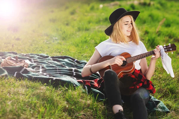 Siyah şapkalı genç ve güzel bir kadının portresi. Çimlerin üzerinde oturan ve gitar çalan kız