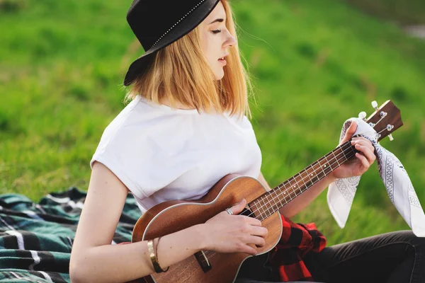 Siyah şapkalı genç ve güzel bir kadının portresi. Çimlerin üzerinde oturan ve gitar çalan kız