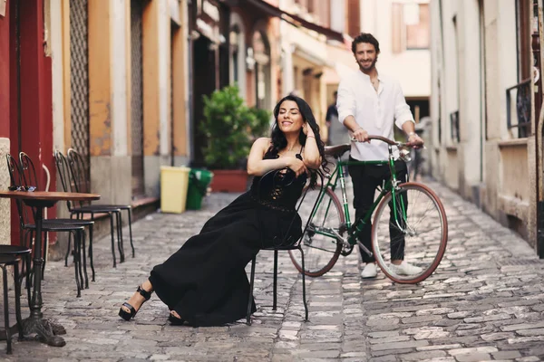 Sepasang kekasih di kota tua. Seorang wanita dengan gaun hitam duduk di kursi. Pria di belakangnya berdiri dengan sepeda hijau Stok Gambar Bebas Royalti