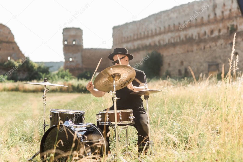 Drummer in black hat rehearses in the open air. He plays drums in the grass near the old castle