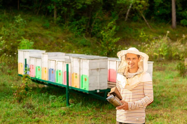 Un apicultor con una tapa protectora prepara una herramienta para fumar abejas de una colmena en el colmenar. Humo para abejas, equipo en colmenar —  Fotos de Stock
