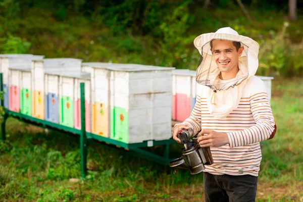 Ένας μελισσοκόμος με προστατευτικό κάλυμμα ετοιμάζει ένα εργαλείο για το κάπνισμα των μελισσών από μια κυψέλη στο μελισσοκομείο. Καπνός για μέλισσες, εξοπλισμός στο μελισσοκομείο — Φωτογραφία Αρχείου