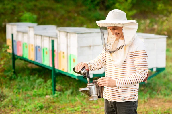 Včelař v ochranném víčku připravuje nástroj pro uzení včel z včelího úlu. Kouř pro včely, vybavení na včelíně — Stock fotografie