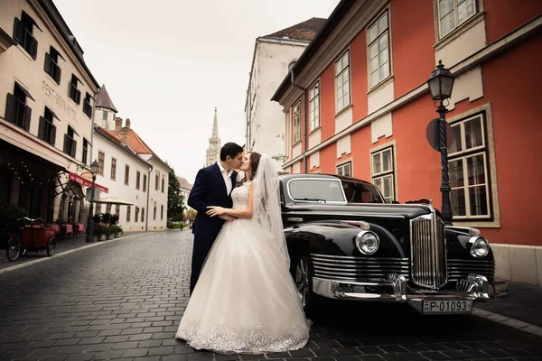 Happy luxury wedding couple kiss and embracing near retro car in old city — Stok Foto