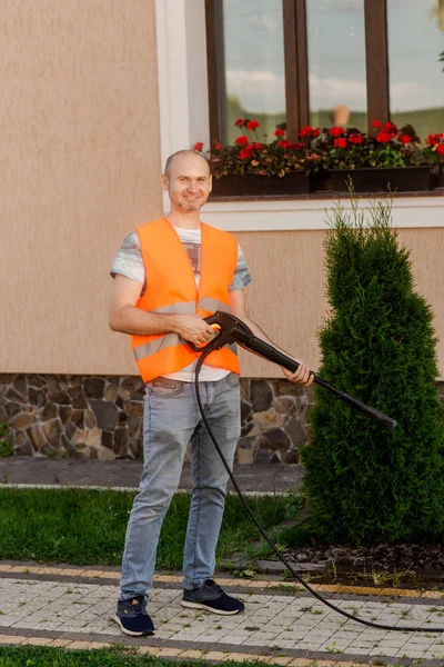 Seorang pria dalam rompi oranye membersihkan genteng rumput di halamannya dekat rumah. Pembersihan tekanan tinggi — Stok Foto