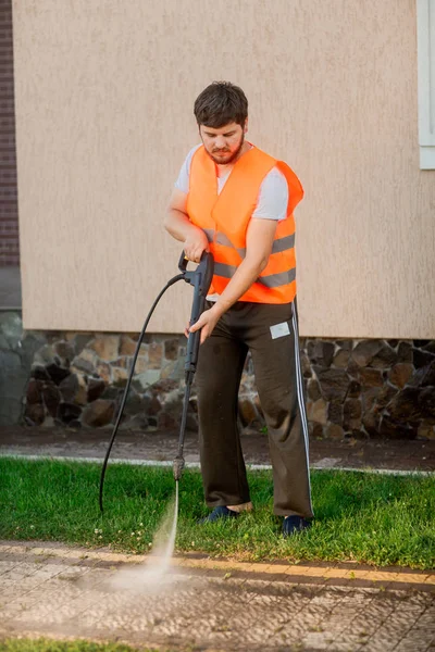Seorang pria dalam rompi oranye membersihkan genteng rumput di halamannya dekat rumah. Pembersihan tekanan tinggi Stok Gambar