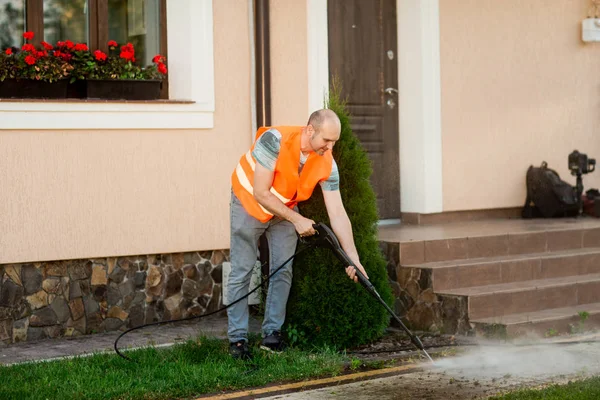 Seorang pria dalam rompi oranye membersihkan genteng rumput di halamannya dekat rumah. Pembersihan tekanan tinggi Stok Foto