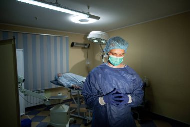 Mavi eldivenli cerrah ameliyatı düşünüyor. Hasta arka plandaki kanepede yatıyor. Ameliyat için hazırlama